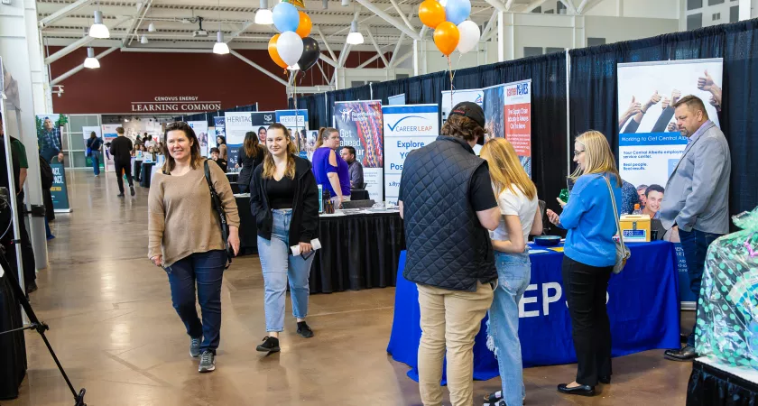 Employer booths line the right side of the photo, small groups of people talking in the booths and two attendees are walking towards the camera.