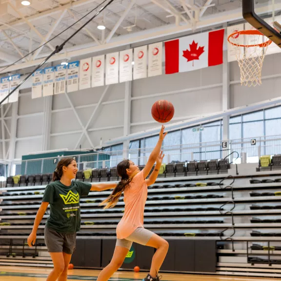 A girl is shooting a basketball towards a hoop while another girl tries to block her